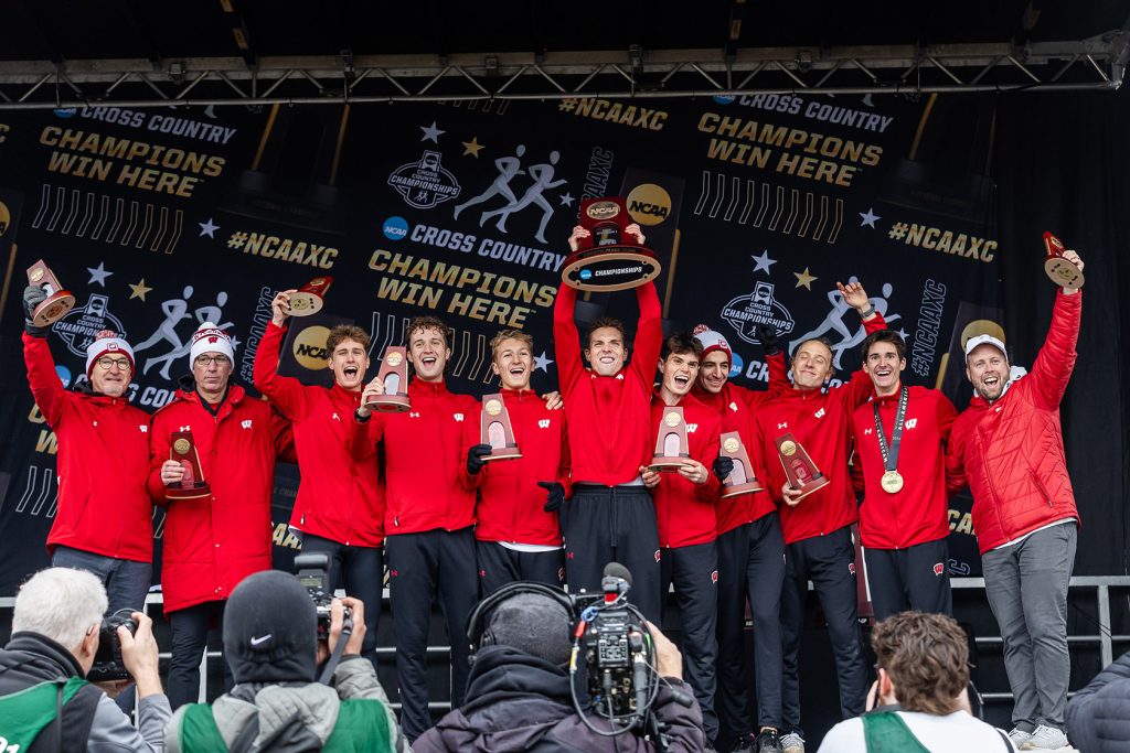 Members of the UW Men's Track team wearing red jackets pose with 2023 NCAA trophies