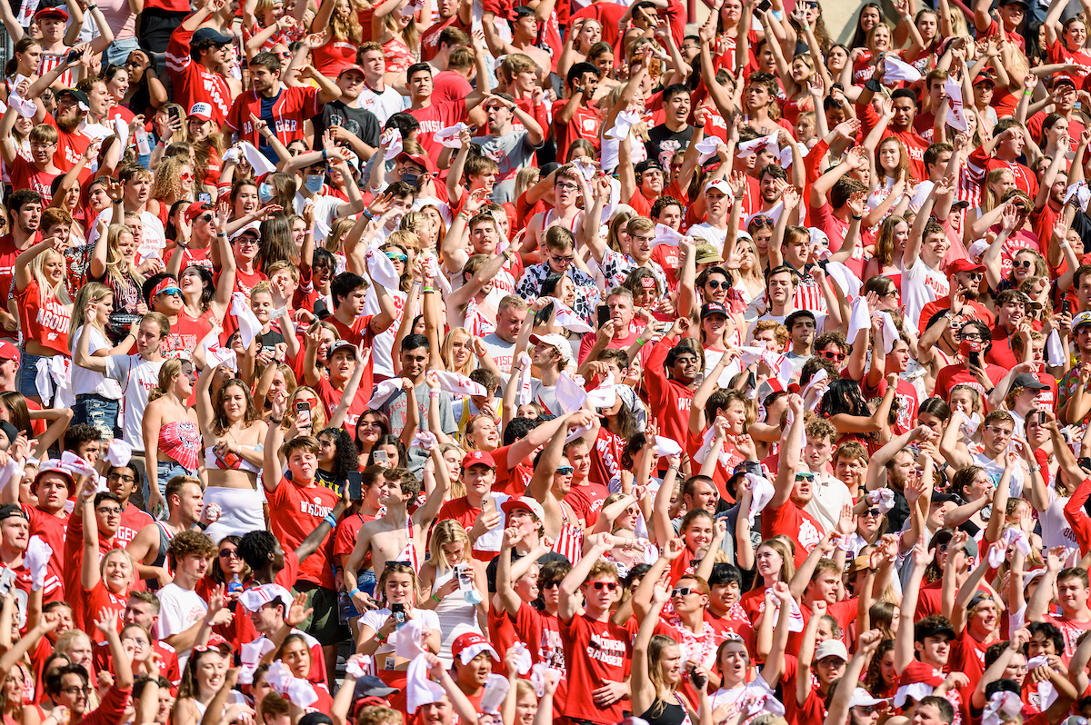 A crowd of people wearing red and white cheer excitedly in the student section at a UW–Madison football game on a sunny day.
