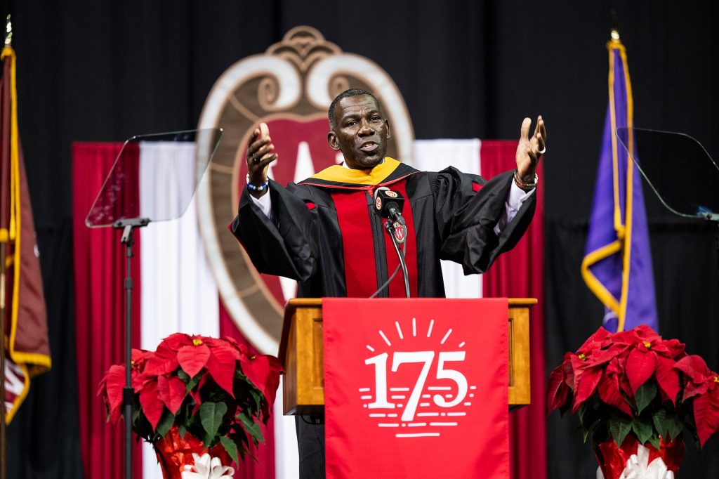 NBA All-Star and UW-Madison alum Michael Finley wearing a Commencement robe gestures while giving the keynote speech during the 2023 Winter Commencement