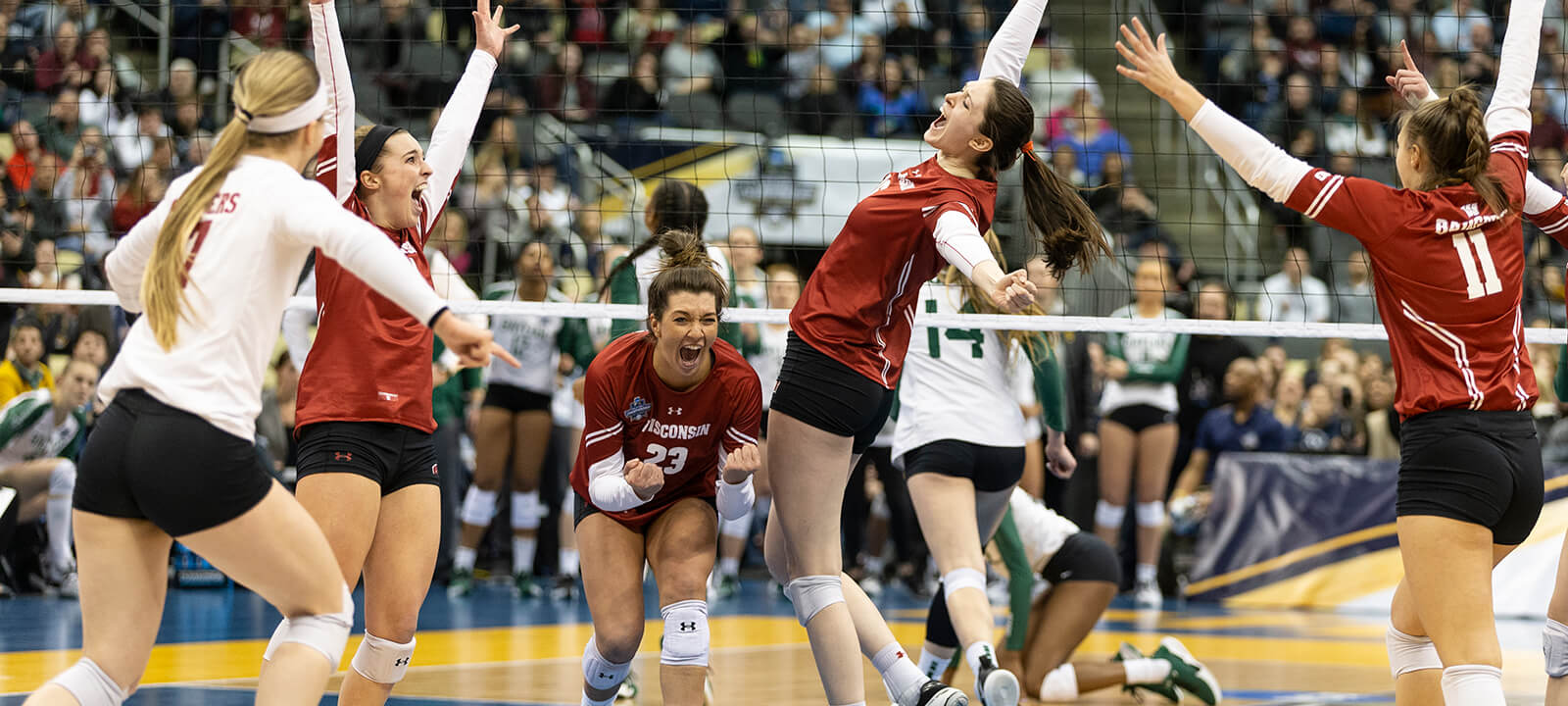 Volleyball players celebrate a win at the net with hands up and smiles.