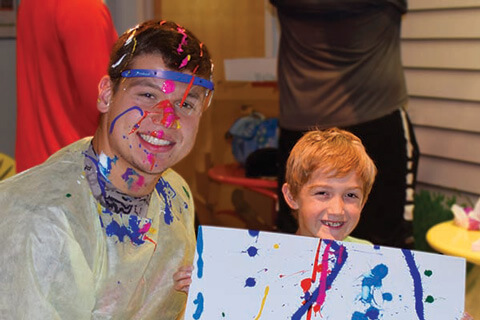 A Badger athlete with paint on his face smiles with a young child holding up a splatter painting project.