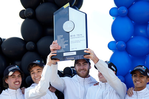 Three smiling members of the men's cross country team hold up an NCAA championship trophy.