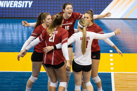 Five Volleyball players celebrate at the net with a group hug and smiles.